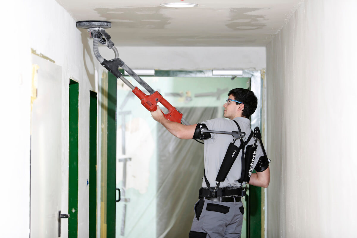 Das Exoskelett Paexo von Ottobock unterstützt Handwerker auf Baustellen bei der Überkopfarbeit, zum Beispiel beim Schleifen der Decke mit einer „Giraffe“ (Teleskopschleifer). (Foto: Ottobock)
