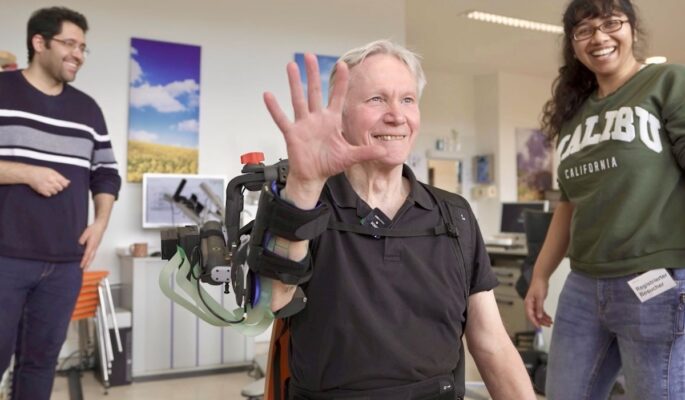 Wolfgang Korisanski ist einer der 24 Patientinnen und Patienten, die das neue System aus Exoskelett und Elektrostimulation für eine Studie bereits getestet haben. Im Hintergrund: Hossein Kavianirad (links) und Neha Das, Forschende aus dem Lehrstuhl von Prof. Sandra Hirche. (Foto: Sabrina Bauer / TUM)